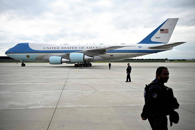 A imagem mostra um avião grande, identificado como o Air Force One, estacionado em uma pista de aeroporto. O avião é predominantemente azul e branco, com a inscrição 'UNITED STATES OF AMERICA' em letras grandes na fuselagem. Há dois homens vestidos com uniformes de segurança próximos ao avião, um deles caminhando e o outro parado. O céu está nublado, e a pista é de concreto