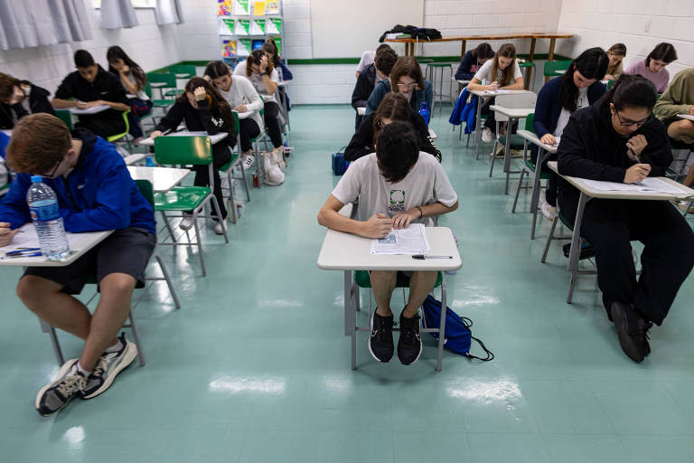 A imagem mostra uma sala de aula com vários alunos realizando uma prova. Os alunos estão sentados em mesas， concentrados em suas atividades. O ambiente é bem iluminado， com janelas ao fundo e algumas paredes decoradas. A maioria dos alunos está vestindo roupas casuais e alguns têm garrafas de água ao lado.