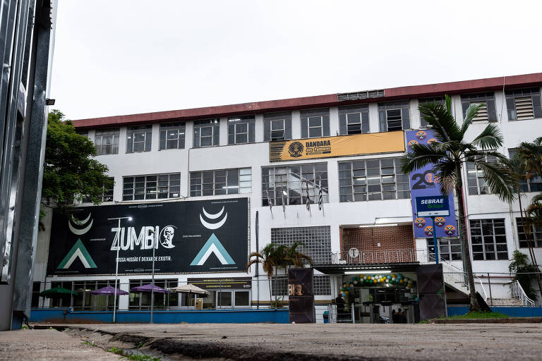 A imagem mostra a fachada de um edifício escolar, identificado como Escola Zumbi dos Palmares. O prédio é de dois andares, com janelas grandes e uma área externa com mesas e cadeiras. Há um grande painel preto com o nome 'ZUMBI' em letras brancas e um símbolo ao lado. Acima, há uma faixa amarela com o nome 'BANDA' e um logotipo. O ambiente é cercado por árvores e plantas.