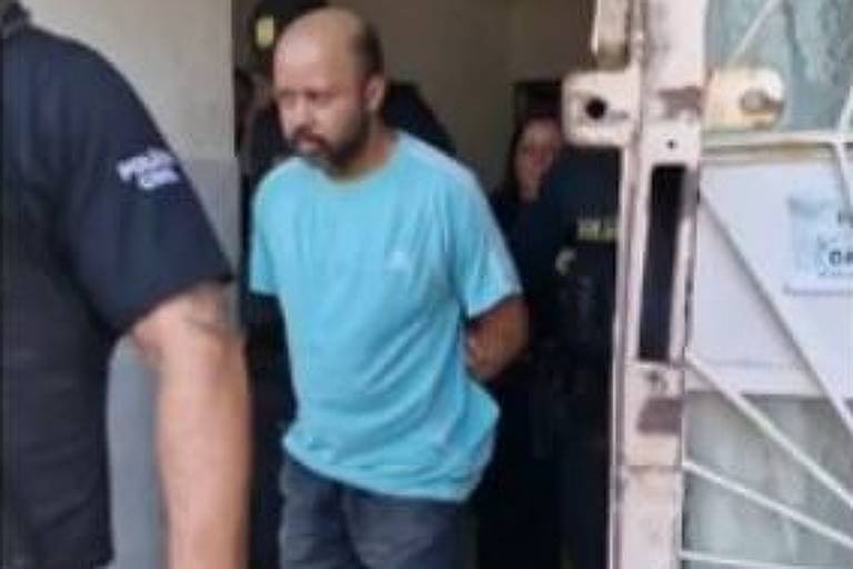 Albino Santos de Lima, homem negro, é conduzido por policias civis. Veste camiseta azul claro, bermuda jeans preta e chinelos de dedo. Está algemado, com as mãos para trás, enquanto caminha saindo de uma residência 