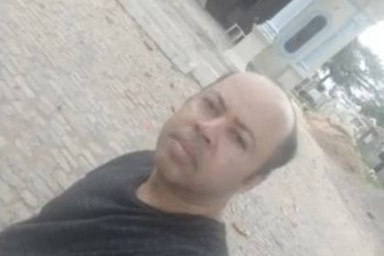 Albino Santos de Lima, homem negro, de camiseta preta, calvo, faz selfie em cemitério. Ao fundo há diversas sepulturas