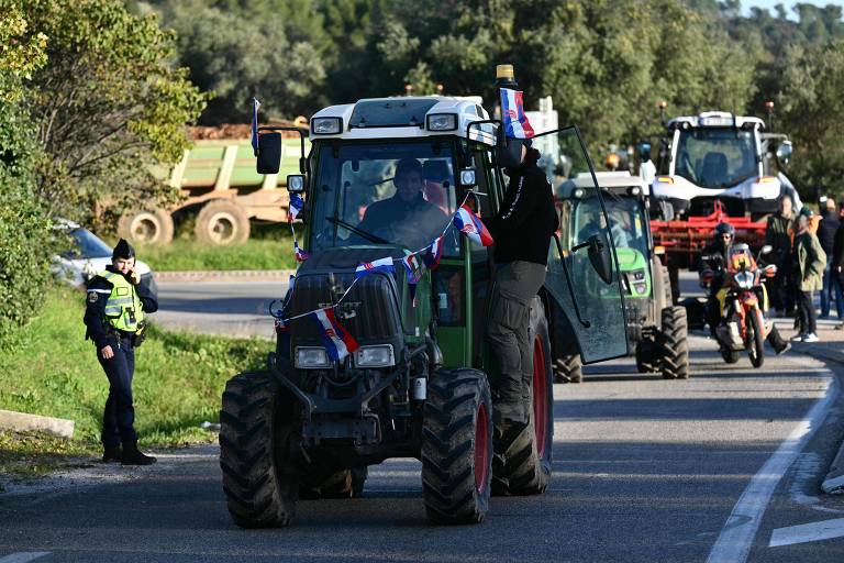 A imagem mostra um cortejo de tratores em uma estrada. Um trator verde está em primeiro plano, decorado com fitas. Um homem está em pé ao lado do trator, enquanto um policial observa. Ao fundo, outros tratores e veículos agrícolas estão alinhados na estrada, cercados por vegetação.