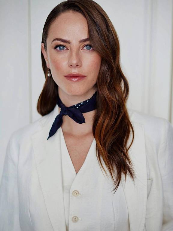 A imagem mostra uma mulher com cabelo castanho claro e ondulado, usando um blazer branco e uma blusa branca. Ela está usando um lenço azul escuro amarrado no pescoço. O fundo é claro, com uma parede branca. A mulher tem um olhar sério e expressivo, com maquiagem suave e brincos pequenos.