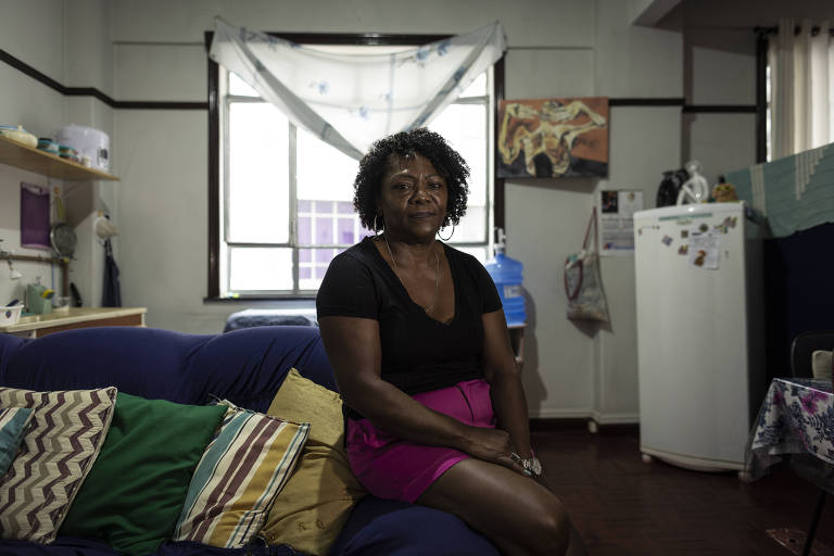 A imagem mostra uma mulher sentada em um sofá azul, cercada por almofadas coloridas. Ela usa uma blusa preta e uma saia rosa. Ao fundo, há uma janela com cortinas brancas e uma parede decorada. O ambiente é uma sala de estar, com uma geladeira branca e prateleiras com objetos diversos.