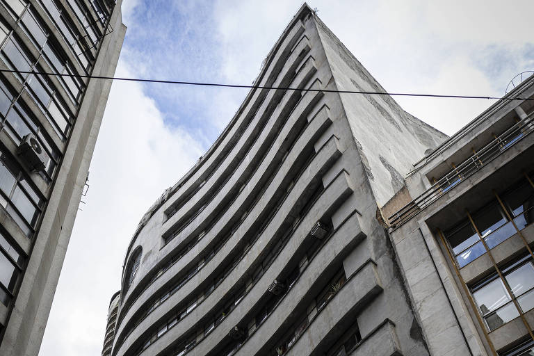 Imagem de um edifício curvo, com várias janelas em sua fachada. O prédio tem um design moderno e é visto de baixo para cima, destacando suas linhas onduladas. O céu está parcialmente nublado ao fundo.