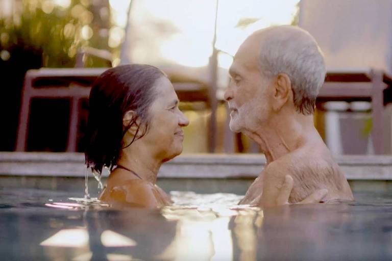 A imagem mostra um casal idoso em uma piscina, olhando um para o outro com sorrisos. A mulher tem cabelo escuro e molhado, enquanto o homem tem cabelo grisalho e uma barba curta. O fundo apresenta uma iluminação suave do pôr do sol, com algumas cadeiras visíveis ao fundo