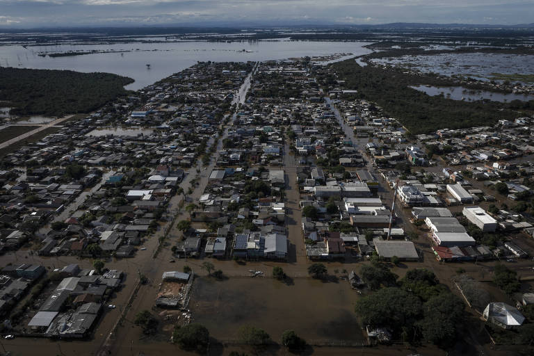 Imagem aérea de uma cidade inundada, mostrando ruas alagadas e casas. O cenário inclui áreas de água visíveis ao fundo, com uma densa concentração de edifícios e vegetação ao redor. O céu está nublado, sugerindo um clima sombrio.