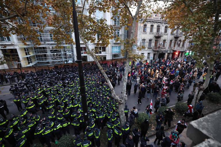 A imagem mostra uma cena de conflito entre manifestantes e a polícia em uma rua. À esquerda, há um grande grupo de policiais em uniformes escuros com coletes amarelos, formando uma linha. À direita, um grupo de manifestantes está reunido, com algumas pessoas segurando cartazes. O ambiente urbano é visível, com prédios e árvores ao fundo, e a iluminação sugere que é ao entardecer.