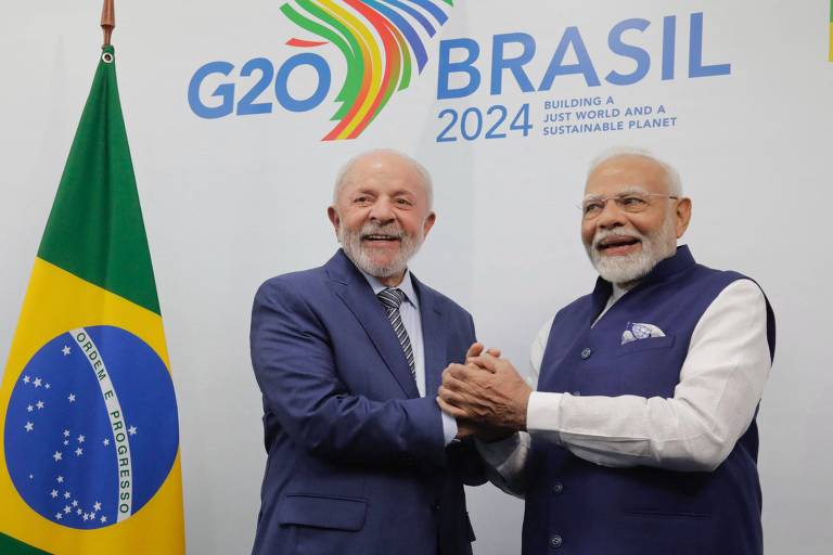 Na imagem, duas pessoas estão se cumprimentando com um aperto de mãos. Ao fundo, há uma bandeira do Brasil e um painel com a inscrição 'G20 BRASIL 2024'.