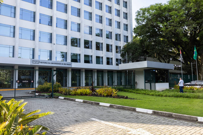 Imagem de um edifício moderno com fachada de vidro e estrutura retangular. Na frente， há um jardim bem cuidado com gramado verde e plantas ornamentais.
