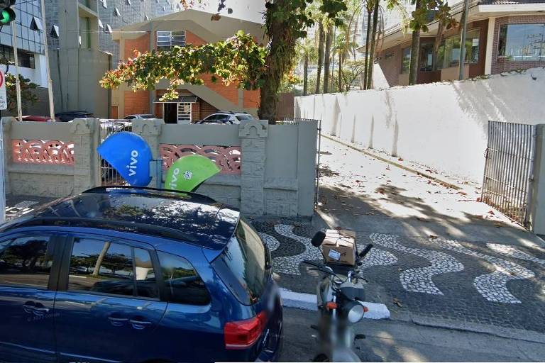 A imagem mostra uma rua urbana com um carro azul estacionado à frente. No fundo， há um prédio de dois andares com uma fachada em tijolos. À esquerda， um sinal de trânsito indica proibição de estacionamento