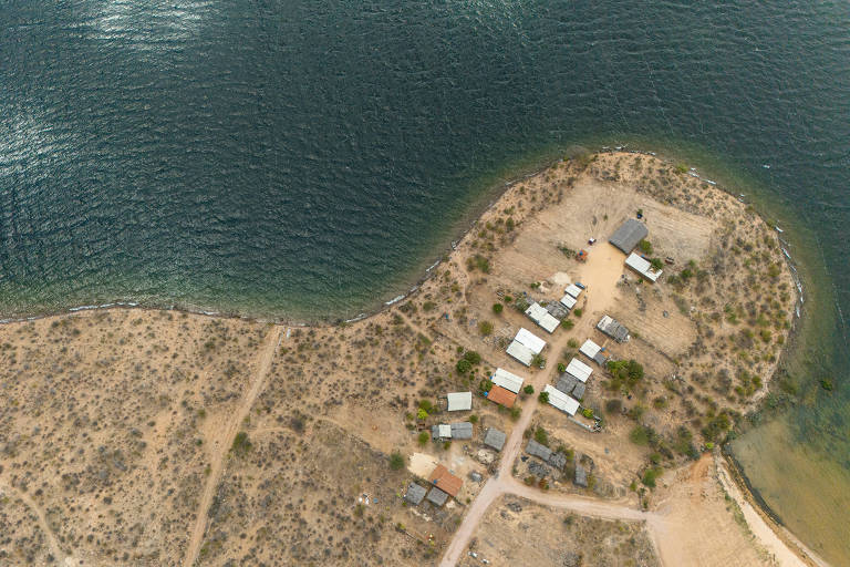 A imagem mostra uma vista aérea de uma pequena península com várias construções, cercada por água. A área é predominantemente árida, com vegetação escassa e um caminho de terra que leva às edificações. O rio apresenta uma coloração azulada e reflete a luz do sol.