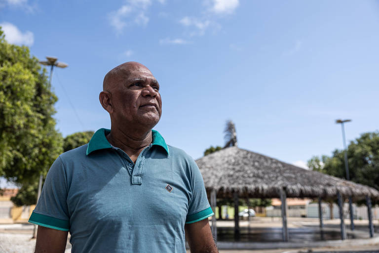 Um homem de pele bronzeada e cabelo raspado está em pé, olhando para o horizonte. Ele usa uma camiseta azul com detalhes em verde. Ao fundo, há uma estrutura coberta com telhado de palha e árvores ao redor, sob um céu claro com algumas nuvens.