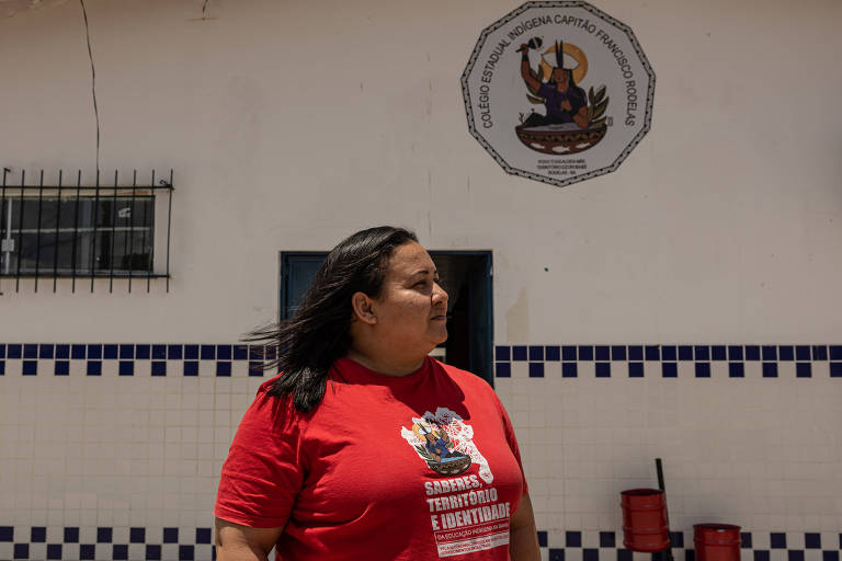 A imagem mostra uma mulher com cabelo longo e solto, vestindo uma camiseta vermelha. Ela está de perfil, olhando para o lado, em frente a uma parede branca com um emblema de uma mulher indígena no topo. A parede tem azulejos azuis na parte inferior. Ao fundo, há uma janela com grades.