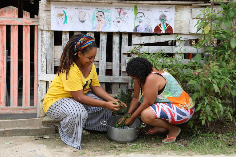 Duas mulheres estão agachadas em um espaço externo, uma delas com uma blusa amarela e calças listradas, e a outra vestindo uma blusa colorida e shorts. Elas estão manipulando plantas em uma bacia. Ao fundo, há uma parede de madeira com desenhos pendurados, representando rostos. O ambiente é natural, com vegetação ao redor.