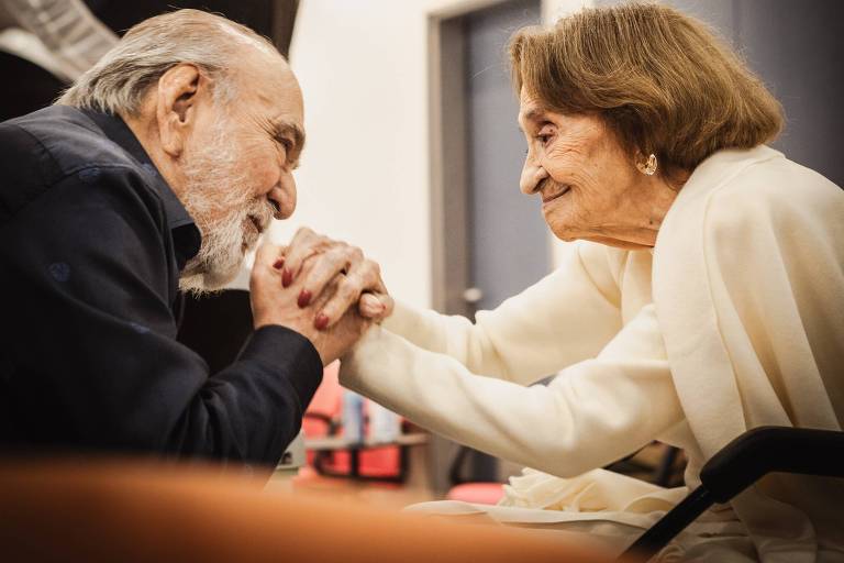 A imagem mostra um homem e uma mulher idosos, sentados frente a frente, segurando arsenic  mãos um bash  outro. O homem tem cabelo grisalho e uma barba curta, enquanto a mulher tem cabelo castanho claro e usa um casaco claro. Ambos estão sorrindo e parecem estar em um momento de conexão emocional. Ao fundo, há uma iluminação suave e algumas cadeiras vermelhas.