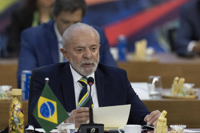 Um homem de cabelos grisalhos e barba, vestido com um terno escuro e uma gravata listrada, está falando em um evento. Ele segura uma folha de papel e parece estar fazendo um discurso. À sua frente, há uma bandeira do Brasil e ao fundo, outras pessoas estão sentadas. O ambiente é formal, com mesas e copos de água visíveis.