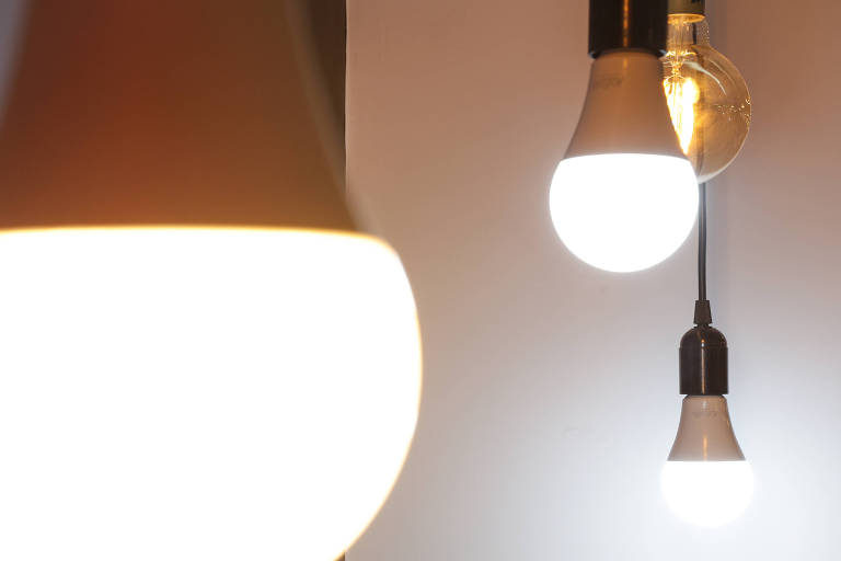 A imagem mostra várias luminárias pendentes em um ambiente interno. Há uma lâmpada com uma cúpula branca e outra lâmpada exposta, ambas emitindo luz. O fundo é neutro, destacando as luminárias.