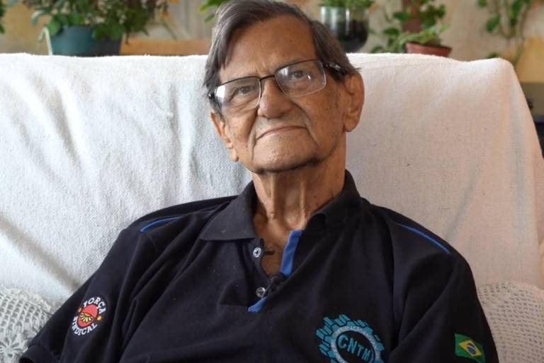 Um homem idoso está sentado em um sofá, usando uma camisa preta com detalhes em azul. Ele tem cabelo grisalho e usa óculos. Ao fundo, há plantas em vasos e uma parede clara.