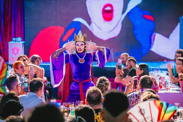 A imagem mostra uma performance drag em um ambiente festivo. Um artista drag, vestido com um traje roxo e uma coroa dourada, está no centro do palco, fazendo uma pose dramática com as mãos. Ao fundo, há uma projeção de uma figura feminina com lábios vermelhos. O público, composto por várias pessoas, está assistindo e registrando o momento com seus celulares. A mesa à frente está cheia de bebidas e pratos.