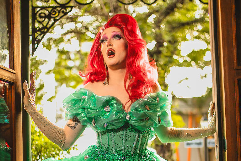 Uma drag queen está em uma entrada de porta, usando um vestido verde volumoso com camadas e detalhes brilhantes. O vestido possui mangas bufantes e é adornado com pedras brilhantes. A drag tem cabelo longo e vermelho, com maquiagem vibrante. O fundo mostra uma área externa com árvores e luz natural.