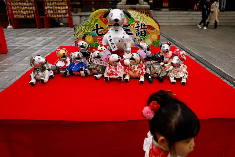 A imagem mostra uma mesa coberta com um tecido vermelho, onde estão dispostos vários bonecos de cachorro. No centro, há um boneco maior vestido com roupas tradicionais, cercado por vários bonecos menores, cada um com trajes coloridos e variados. Ao fundo, pode-se ver uma decoração festiva. Uma criança com cabelo preso em um coque e um laço rosa aparece na parte inferior da imagem, parcialmente visível.