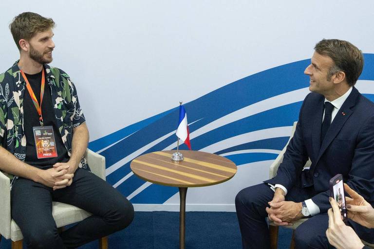 A imagem mostra um encontro durante o G20 Brasil 2024. À esquerda, um homem com cabelo curto e barba, vestindo uma camisa estampada e calças escuras, está sentado em uma cadeira. À direita, um homem com cabelo curto e bem vestido, usando um terno escuro, também está sentado. Entre eles, há uma mesa redonda com uma bandeira da França. Ao fundo, está o logotipo do G20 Brasil 2024, com as palavras 'G20 RIO SUMMIT' e 'PRÉSIDENT' em destaque.