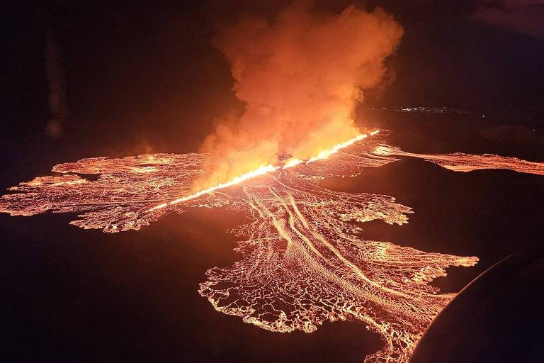 A imagem mostra uma erupção vulcânica à noite, com lava fluindo em várias direções. A lava brilha intensamente em tons de laranja e vermelho, enquanto uma densa coluna de fumaça se eleva no ar. O cenário é iluminado pela luz da lava, destacando o terreno ao redor.