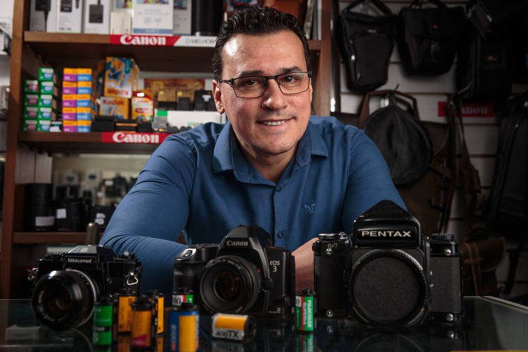 Um homem branco sorridente， de cabelos curtos ondulados， com camisa azul de tom escuro e óculos retangulares de armação preta， está posicionado em frente a uma mesa com três câmeras fotográficas: uma Nikkormat à esquerda， uma Canon EOS 3 no centro e uma Pentax à direita. Na mesa， há também várias bobinas de filme. Ao fundo， prateleiras com produtos relacionados à fotografia， incluindo caixas de filmes Kodak e outros acessórios. 