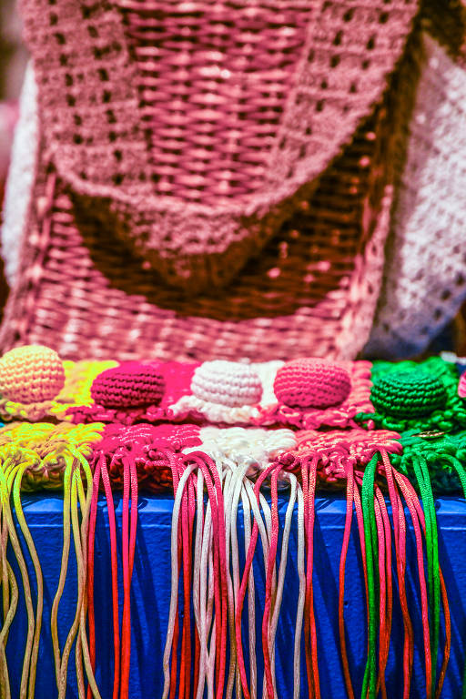 A imagem mostra uma mesa com várias bolas de lã coloridas, incluindo rosa, branco, amarelo, verde e azul. Ao fundo, há uma peça de crochê em tons de rosa, que parece ser uma bolsa ou um cachecol. As linhas de lã estão dispostas de forma organizada, criando um contraste vibrante com o fundo.