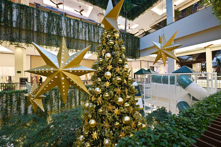 A imagem mostra uma árvore de Natal decorada com estrelas douradas e bolas brilhantes em um shopping. Ao fundo, há um espaço com vegetação e um escorregador em uma área de recreação. Estrelas grandes e douradas estão penduradas no teto, complementando a decoração festiva.