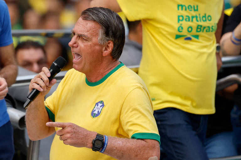 Bolsonaro é um homem com camiseta amarela, que possui a bandeira do Brasil e está falando ao microfone em um evento. Ele parece estar animado e gesticulando com a mão. Ao fundo, há pessoas vestindo camisetas amarelas e uma delas tem uma frase em verde que diz 'meu partido é o Brasil'.