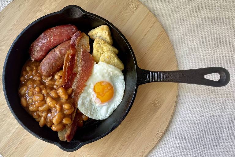 Uma frigideira de ferro fundido contém um café da manhã que inclui salsichas， bacon， feijão， um ovo frito com gema amarela e pedaços de batata. O prato está sobre uma superfície de madeira clara.