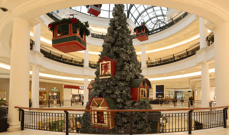 A imagem mostra uma grande árvore de Natal decorada com luzes e enfeites, localizada no centro de um shopping. A árvore possui casas decorativas em sua base e está cercada por um corrimão. Acima da árvore, há cabines de teleférico decoradas com folhagens e laços vermelhos. O ambiente é iluminado e apresenta lojas ao fundo, com uma arquitetura moderna e elegante.