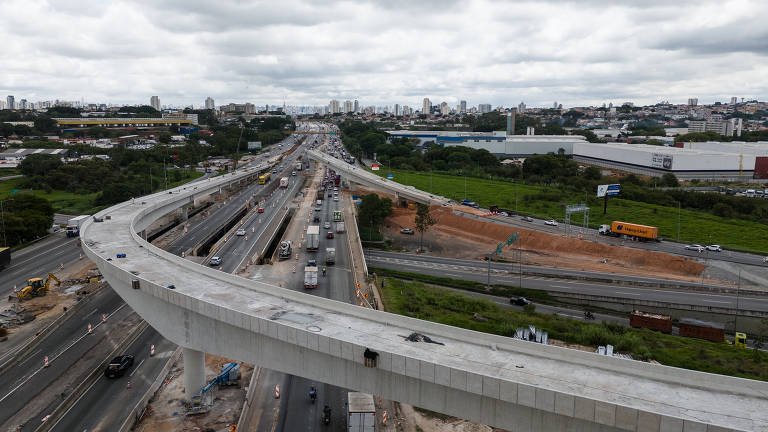 A imagem mostra uma vista aérea de uma rodovia em construção, com várias faixas de tráfego visíveis. À esquerda, há uma estrutura elevada em concreto, enquanto à direita, a rodovia está em obras, com máquinas e materiais de construção. Ao fundo, pode-se ver a cidade com prédios e uma cobertura de nuvens no céu