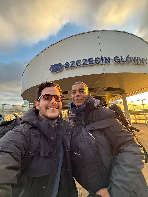 A imagem mostra duas pessoas tirando uma selfie em frente à estação de trem Szczecin Główny. Ambas estão usando jaquetas escuras e carregam mochilas. O céu ao fundo apresenta nuvens e um tom dourado， indicando o pôr do sol.