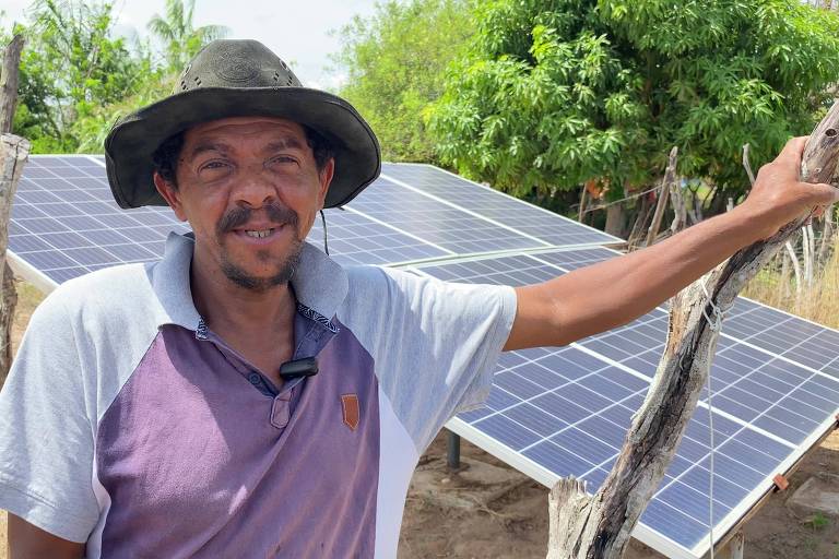 Um homem está posando ao lado de um conjunto de painéis solares. Ele usa um chapéu e uma camisa de manga curta, sorrindo para a câmera. Ao fundo, há vegetação e árvores, indicando um ambiente rural.