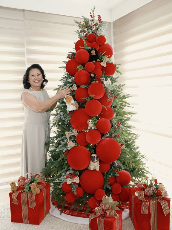 A imagem mostra uma mulher sorridente ao lado de uma árvore de Natal decorada com bolas vermelhas grandes e pequenos enfeites. A árvore está adornada com detalhes verdes e algumas figuras de animais. Ao redor da árvore, há presentes embrulhados em papel vermelho com laços coloridos. O ambiente é iluminado e possui cortinas claras ao fundo.