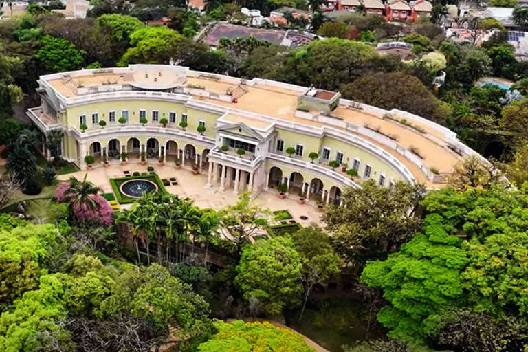 A imagem mostra uma mansão em formato curvo, cercada por uma densa vegetação verde. A estrutura possui colunas na entrada e janelas com vasos de plantas. No centro do jardim, há um pequeno lago circular. O telhado da mansão é plano e a área ao redor é repleta de árvores e arbustos, com algumas flores visíveis.