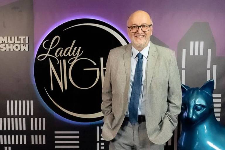 Um homem de cabelos grisalhos e barba, usando um terno cinza claro e uma gravata azul, está posando em frente a um fundo que apresenta o logotipo do programa 'Lady Night'. Ao lado dele, há uma escultura de um gato azul. O fundo é decorado com silhuetas de prédios e o texto 'MULTI SHOW' está visível no canto superior esquerdo.