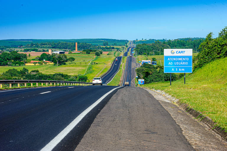 A imagem mostra uma estrada ampla e reta que se estende em direção ao horizonte, cercada por campos verdes e árvores. À direita, há um painel de sinalização azul com texto visível. O céu está limpo e azul, e alguns veículos podem ser vistos na estrada