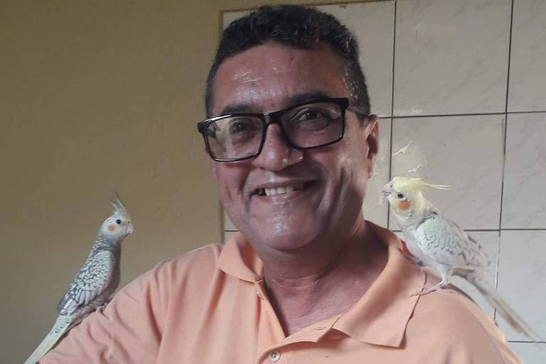 Um homem sorridente está posando para a foto com duas aves brancas em seus ombros. Ele usa uma camisa polo laranja e óculos. O fundo é de uma parede clara, e o ambiente parece ser interno.