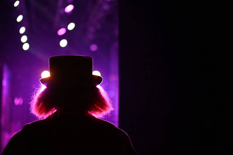 A imagem mostra a silhueta de uma pessoa com cabelo volumoso e um chapéu, vista de costas. Ao fundo, há luzes coloridas em tons de roxo e branco, criando um efeito dramático. A cena sugere um ambiente de apresentação ou espetáculo.