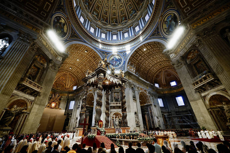 A imagem mostra o interior da Basílica de São Pedro, com uma grande cúpula ornamentada e detalhes arquitetônicos elaborados. No centro, há um altar elevado, cercado por colunas e estátuas. A iluminação destaca a grandiosidade do espaço, enquanto uma multidão de pessoas está reunida em frente ao altar, participando de uma cerimônia religiosa.