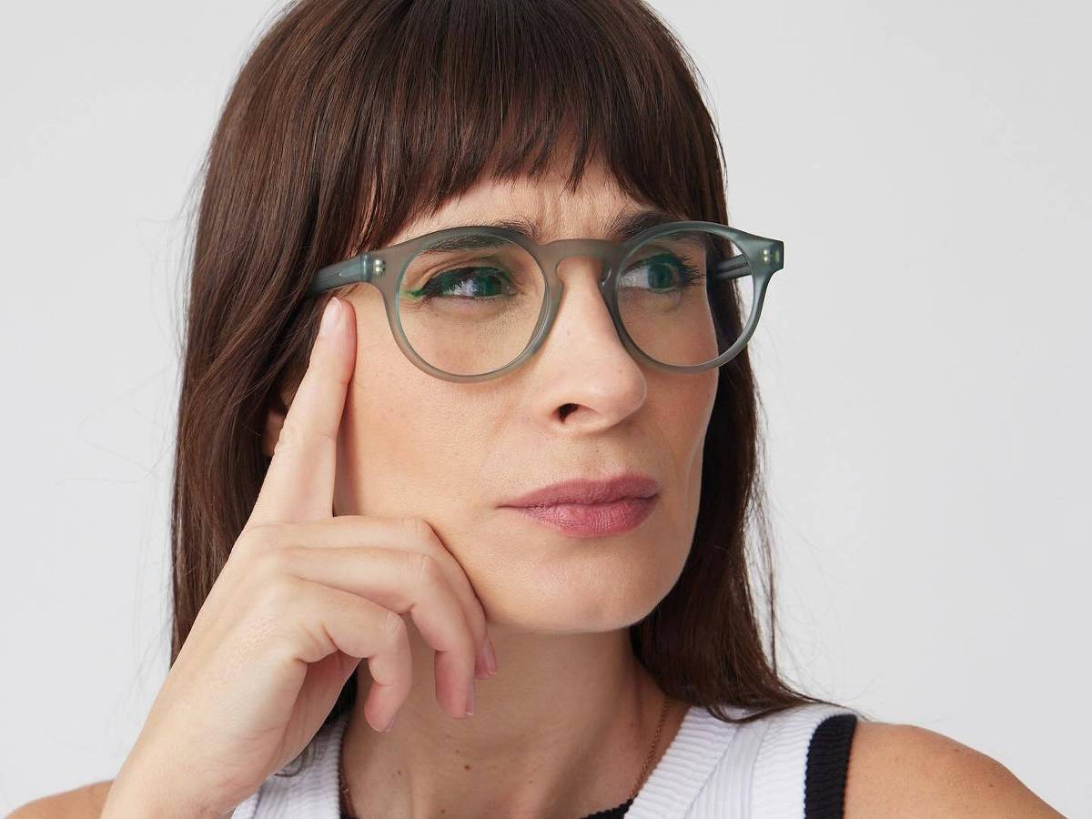 Camila Fremder *** Local Caption *** A imagem mostra uma mulher com cabelo castanho escuro e franja, usando óculos de armação verde. Ela está com a mão direita encostada no rosto, em uma pose pensativa. A mulher veste uma blusa sem mangas, com um detalhe em preto na parte superior. O fundo é branco.