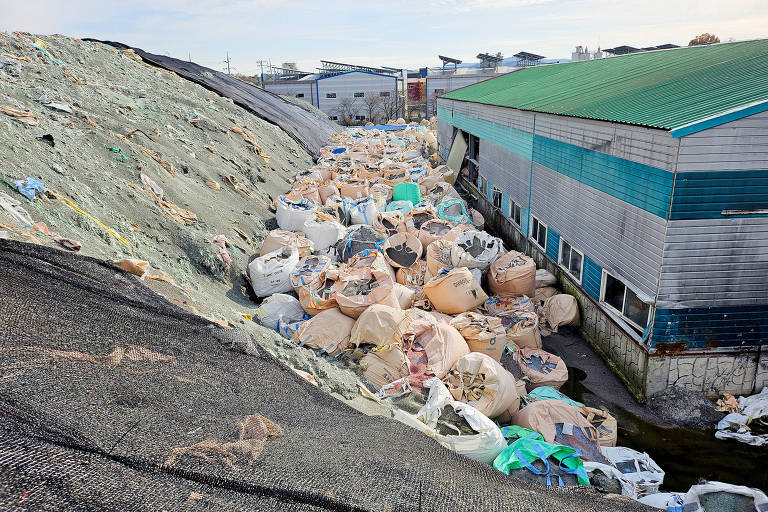 A imagem mostra uma área de descarte de resíduos, onde grandes sacos de lixo estão empilhados ao longo de uma encosta. Ao fundo, há um edifício com telhado verde e paredes de metal, parcialmente coberto por resíduos. O ambiente parece desordenado, com uma grande quantidade de materiais acumulados.
