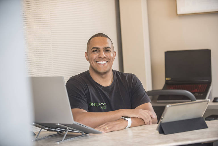 Um homem sorridente， de cabelo curto， está sentado em uma mesa de trabalho. Ele usa uma camiseta preta com a inscrição 039;ONCARD DIGITAL039; em verde e tem um relógio de cor clar no pulso esquerdo. À sua frente， há um laptop e um tablet em um suporte. O ambiente é iluminado e moderno， com uma parede clara ao fundo.