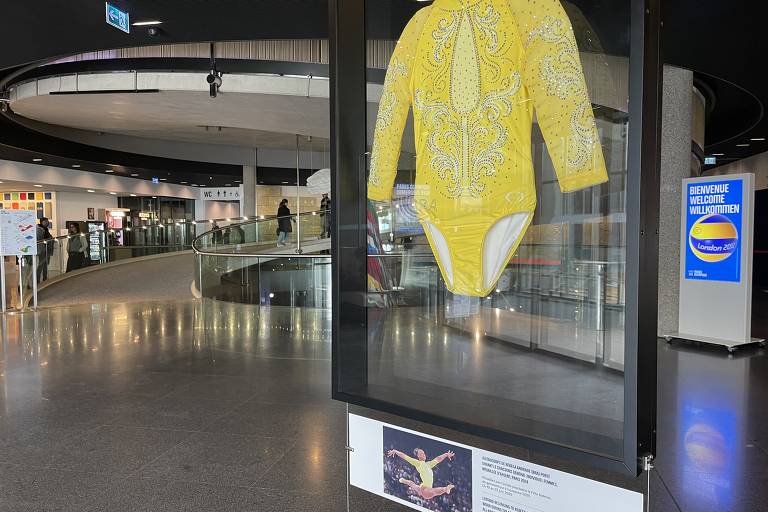 A imagem mostra um traje de banho amarelo exposto em uma vitrine em um espaço de exposição. O traje é de manga longa e possui um design simples. Ao fundo, há uma área ampla com pessoas e uma escada rolante, além de painéis informativos e iluminação suave.