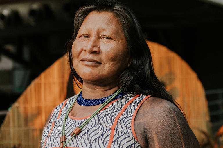 A imagem mostra uma mulher indígena sorrindo, com cabelo longo e solto. Ela está vestindo uma blusa com padrões geométricos e colares. Ao fundo, há uma decoração natural com plantas e uma iluminação suave.
