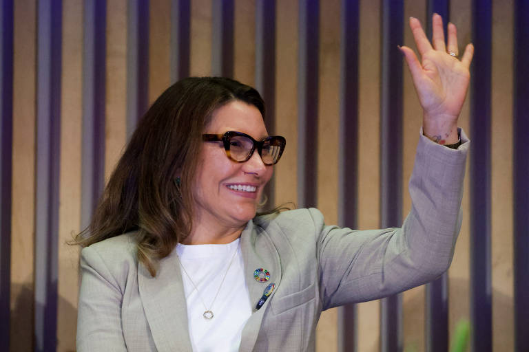 Janja é uma mulher com cabelo castanho claro e ondulado, usando óculos grandes e uma jaqueta cinza. A primeira-dama está sorrindo e acenando com a mão direita. Ela parece estar em um evento, com um fundo de painéis de madeira e algumas plantas visíveis.